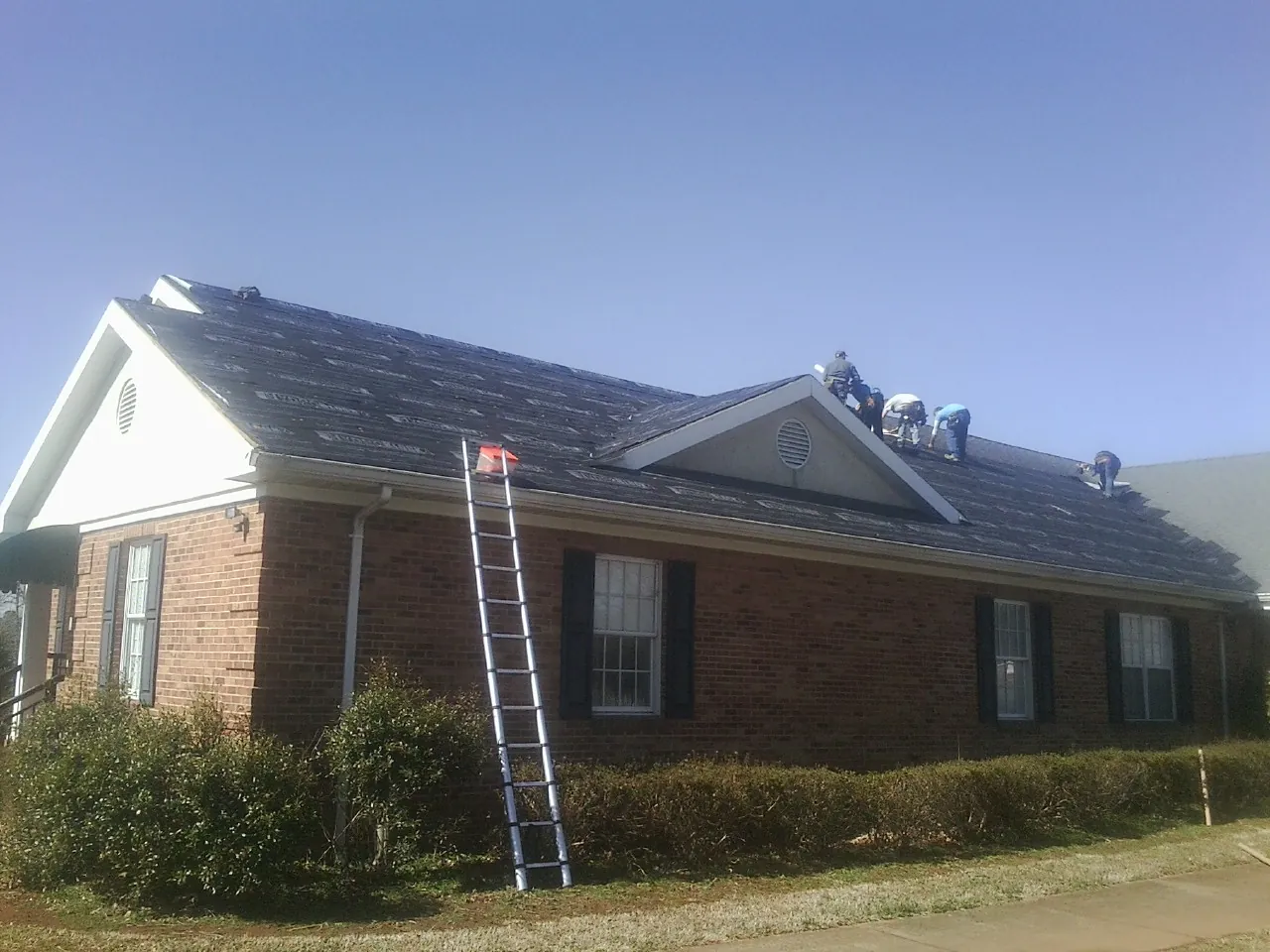 Crew tearing shingles off a brick ranch roof with a ladder set — Metal Roof Repair work by our crew in Detroit Lakes