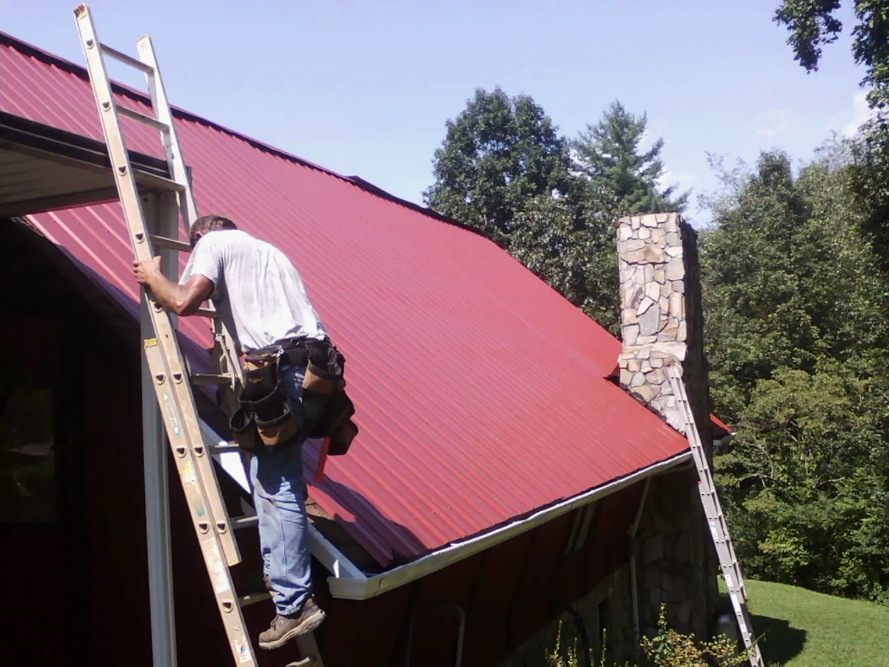Roofer on a ladder beside a red metal roof and stone chimney — Soffit & Fascia Repair by Roofing Brothers in Detroit Lakes
